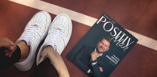 Magazín POSITIV MAN s Romanem Valným na titulce položený na sportovní ploše vedle bílých tenisek – spojení stylu, vůle a inspirace.